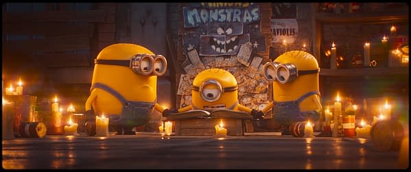 Three Minions gathered around a book, surrounded by glowing candles, with a mysterious wall background featuring a sign that reads "Minions & Monstruos." The scene has a whimsical and slightly spooky atmosphere.