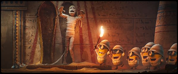 A cartoon scene depicts a mummy standing in an ancient tomb while several excited Minions in explorer outfits cheer and hold a torch. The backdrop features hieroglyphics and decorations typical of an Egyptian setting.