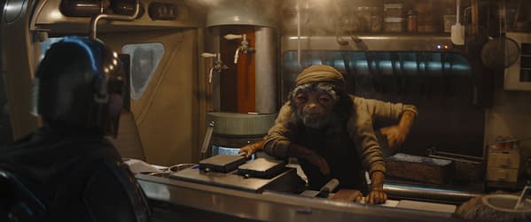 A scene from 'The Mandalorian' featuring the Mandalorian in armor, seated at a counter across from a Fry Cook, who has a furry face and is wearing a cap. The setting appears to be a busy kitchen with steam and cooking equipment in the background.