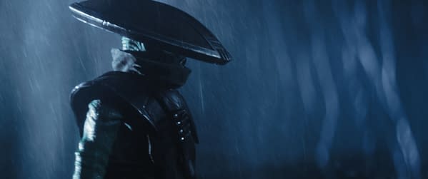 A character dressed as a bounty hunter from Lucasfilm's "The Mandalorian and Grogu" stands in the rain, slightly turned away, with a large distinctive headpiece. The scene conveys a mysterious and atmospheric vibe.