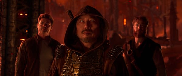 A scene from Mortal Kombat II featuring three characters, with a focus on the central figure wearing a hood and an ornate chestpiece, looking determined against a dark, fiery background. The other two characters stand behind him, contributing to the intense atmosphere.