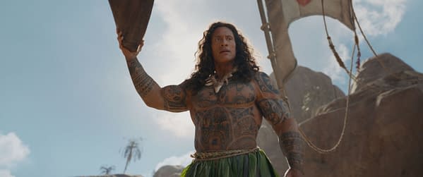 Dwayne Johnson as Maui in the live-action adaptation of Moana, displaying intricate tattoos and holding a wooden oar, with rocky terrain and a clear blue sky in the background.