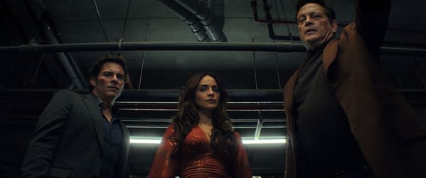 A scene from the film 'Mike & Nick & Nick & Alice' featuring three characters: a man in a gray suit, a woman in a red dress, and an older man in a brown coat, all looking downwards. The setting appears to be an industrial space with visible pipes and overhead lighting.