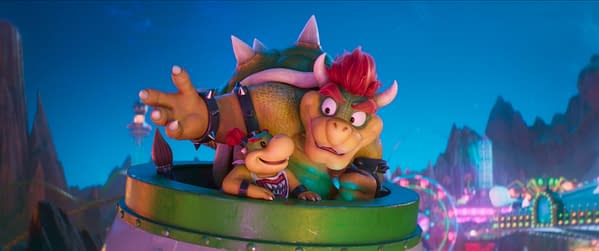 A scene from the Super Mario Movie featuring two characters that resemble Bowser and a smaller version of him. They are in a green vehicle set against a colorful backdrop of mountains and lights, indicating a whimsical, animated atmosphere.