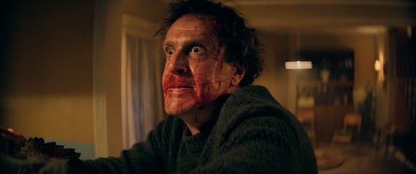A still from the film 'Over Your Dead Body' featuring a man with a distressed expression, covered in blood, wearing a dark sweater, and appearing to be in a tense situation inside a dimly lit room.