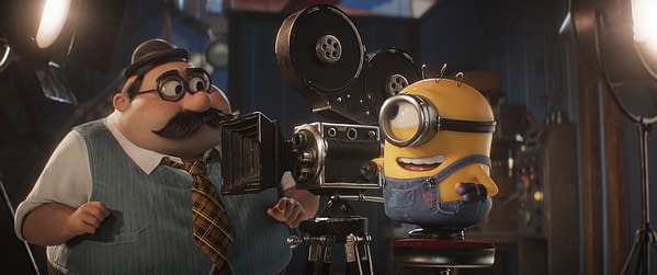 A Minion sits on a vintage film camera while a mustached man in a hat looks on in the studio, from Illumination's Minions & Monsters.