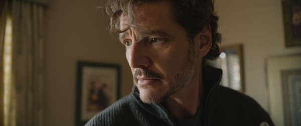 Pedro Pascal in a tense close-up, looking off to the side in warm indoor light. He wears a dark zip-up sweater, with a blurred room in the background.