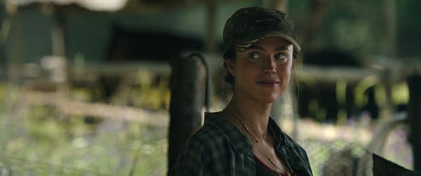 Margaret Qualley as Cima in The Dog Stars, wearing a cap and plaid shirt in a dimly lit outdoor setting.