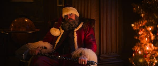 David Harbour as Santa Claus sits in a dimly lit room in Violent Night 2, wearing a red velvet suit and holding a cane. A glowing Christmas tree blurs in the foreground.