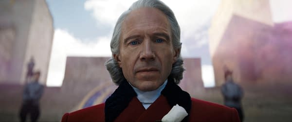 Ralph Fiennes as President Snow stands in a red military-style coat in The Hunger Games: Sunrise on the Reaping. Blurred figures and a bright outdoor backdrop loom behind him.