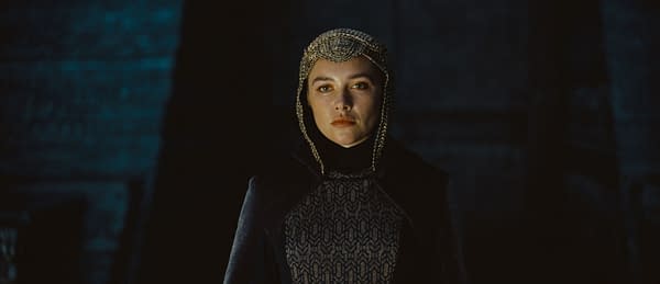 A character from 'Dune: Part Three' stands confidently, dressed in a dark, elaborately patterned outfit with a decorative headpiece, set against a dimly lit background.