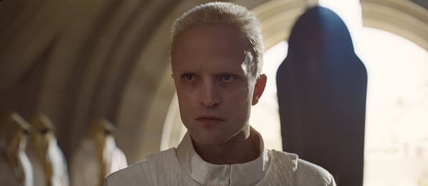 A character from 'Dune: Part Three' with a pale appearance, short blonde hair, and a serious expression, set against a softly lit, architectural background.