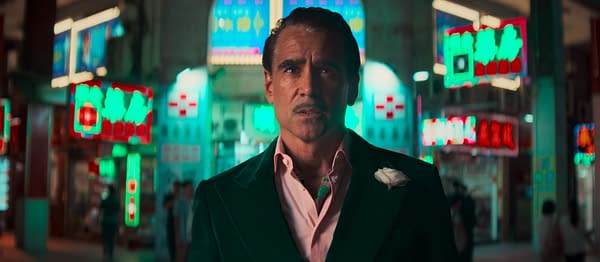 A man with slicked-back hair, dressed in a dark green suit, stands in a brightly lit urban setting filled with colorful neon signs. His serious expression suggests tension, contrasting with the vibrant backdrop.