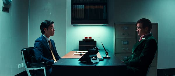 A tense scene in an office setting, featuring two men seated across from each other at a desk. One man in a suit appears to be making a serious point, while the other, dressed in a green suit, listens intently in a well-lit, modern environment.