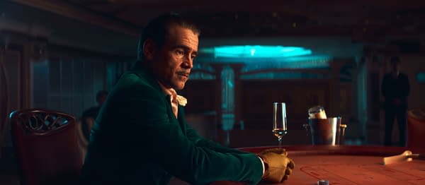 A man with slicked-back hair sits at a poker table, wearing a green blazer and gloves, with a glass of champagne in front of him. The scene is set in a dimly lit casino, creating an atmosphere of tension and intrigue.