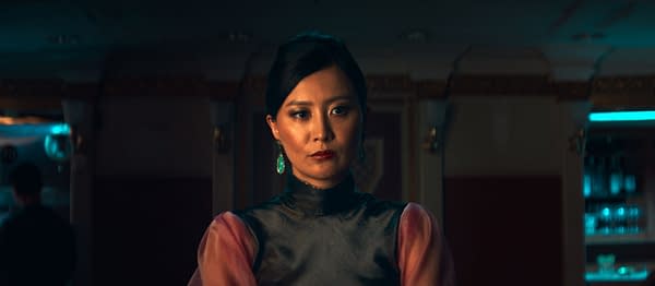 A woman poses in a dimly lit setting, wearing a sleek black outfit with puffed pink sleeves and striking green earrings. Her expression is serious, and the background features soft blue lighting and blurred figures suggesting a social atmosphere.
