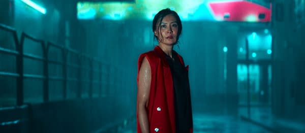 A woman stands confidently in a rainy, neon-lit urban setting, wearing a red sleeveless coat over a black outfit. The wet atmosphere and glowing lights create a dramatic and moody backdrop.