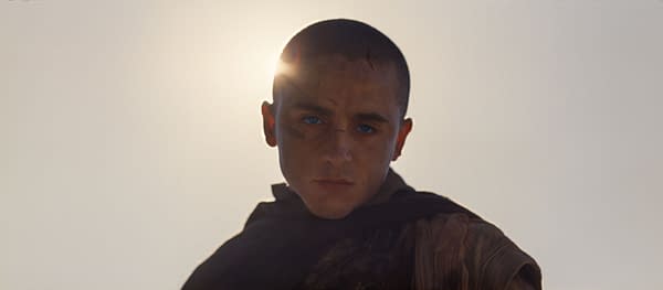A character from 'Dune: Part Three' stares intensely at the viewer, with a bright light source behind them creating a halo effect. The character has short hair, striking blue eyes, and facial markings, dressed in a dark cloak.