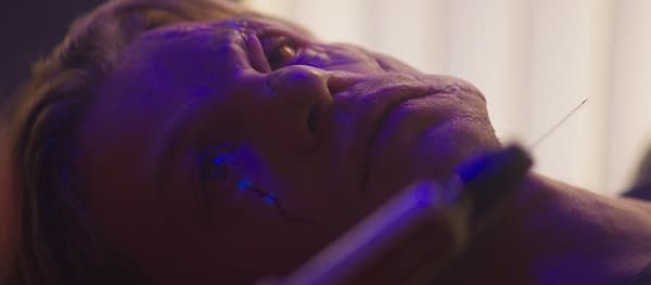 Close-up of a person lying down as a needle approaches their skin, lit with purple-blue light during a tattoo or body modification procedure.