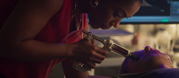 A woman in red uses a syringe-like applicator to apply a treatment to another person's face in a dimly lit medical or cosmetic setting.