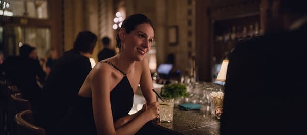 Eva Victor smiles at a dimly lit bar in BEHEMOTH!, wearing a black dress and leaning on the counter with a drink.