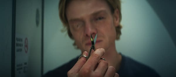 Close-up of a blurred man holding a small pair of scissors in the foreground, with a clinical or industrial setting in the background.