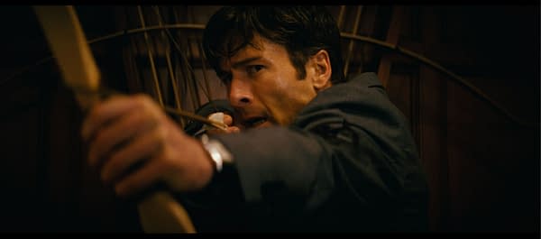 A man in a suit aims an arrow from a bow, displaying intense focus and determination. The background features wooden paneling, adding to the dramatic atmosphere.