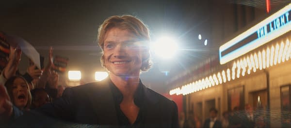 A man with wavy blonde hair smiles brightly in a bustling event, surrounded by excited fans holding up signs in a well-lit atmosphere, reminiscent of a film premiere.