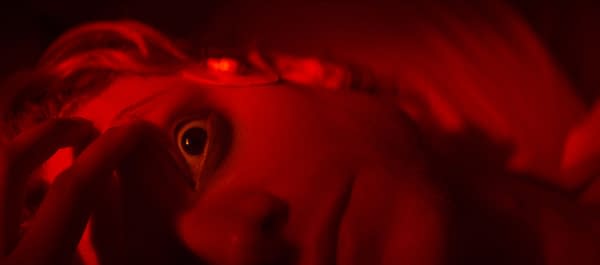 Close-up of a person's face lit in deep red, with one eye visible and fingers near the eye.