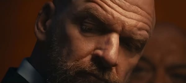 Close-up of a man with a beard and intense expression, wearing a suit. The background features a blurred figure, contributing to a dramatic cinematic moment.