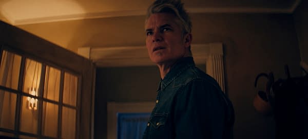 A still from 'Over Your Dead Body' featuring a man with light hair and a denim shirt, looking concerned or startled. The setting is indoors with warm lighting and partially illuminated windows, suggesting a tense atmosphere.