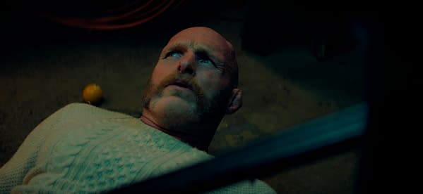 A still from the film 'Over Your Dead Body', featuring a man with a bald head and mustache lying on the ground, looking upward with a concerned expression. An orange is seen in the background, contributing to a tense and suspenseful atmosphere.