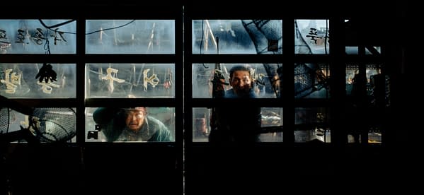 A gritty film still showing two people seen through a weathered, fogged-up window, with one person looking directly out.