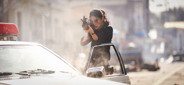 Action still from Hope showing a woman aiming a rifle from beside a police car in a dusty street.