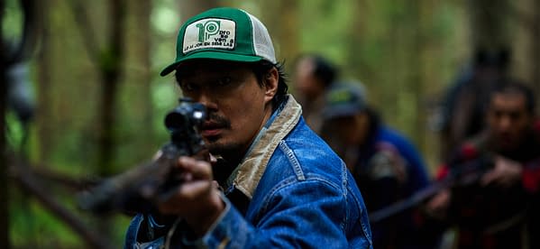 A tense man in a green cap and denim jacket aims a rifle in a forest, with several blurred people behind him.