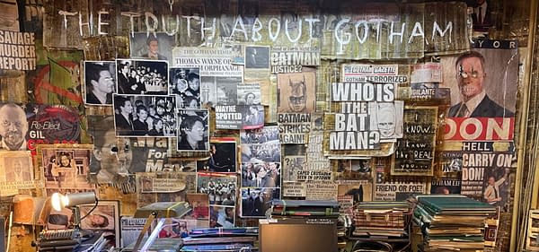 A cluttered desk adorned with piles of books and papers, covered with newspaper clippings about Gotham and Batman. The wall behind features headlines and photos, including questions about Batman's identity, creating a chaotic yet intriguing atmosphere.