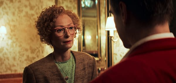 A woman with curly hair and pink glasses engages in a serious conversation with a man, who is partially out of frame. The setting features a patterned wallpaper and elegant lighting.