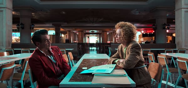 Ballad of a Small Player A still from a film showing two characters in conversation at an empty diner table. The man, dressed in a red blazer, has an intense expression, while the woman, in a beige outfit with curly hair, appears contemplative. Papers are spread out on the table, indicating a serious discussion.