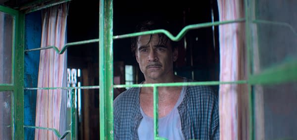 A man stands at a window, looking pensively outside. He has a serious expression, and sweat is visible on his face, suggesting an intense or emotional moment.