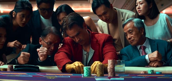 Ballad of a Small Player A group of people, including a man in a red jacket, intensely watch a card game at a casino table, with poker chips and drinks visible. The atmosphere is tense as they focus on the player's next move.