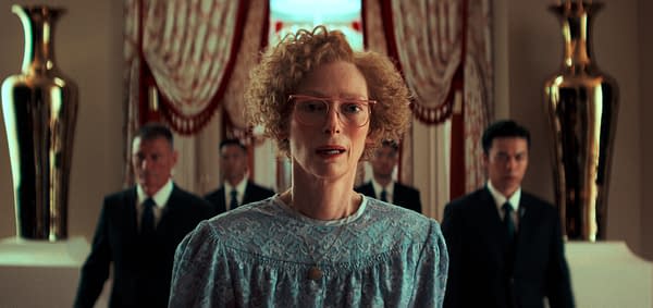A woman with curly hair and glasses stands in the foreground, wearing a blue lace dress, while several men in suits are blurred in the background, creating a tense atmosphere.