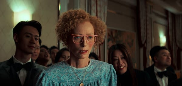 A woman with curly red hair and pink glasses looks intensely surprised or worried in a crowded, elegantly decorated room. People in formal attire surround her, displaying a range of expressions.