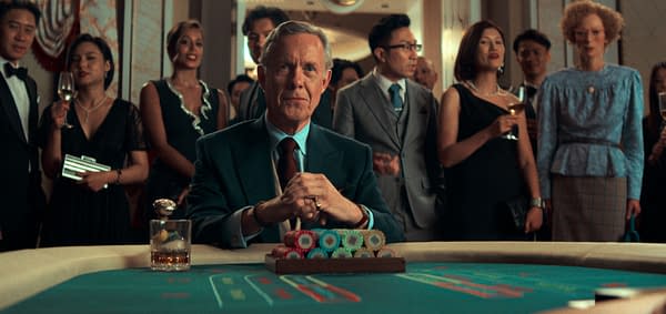 A well-dressed man sits at a casino table, surrounded by a crowd of elegantly dressed people who are watching him intently. He has a glass of whiskey in front of him and a stack of colorful poker chips on the table.