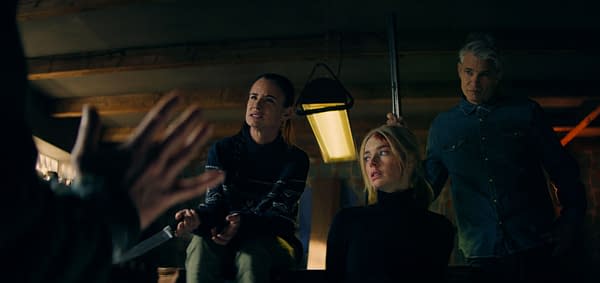 A tense scene from the film 'Over Your Dead Body,' featuring three characters in a dimly lit room. One character appears to be pleading with another, who holds a weapon, while a third character looks on with a worried expression.