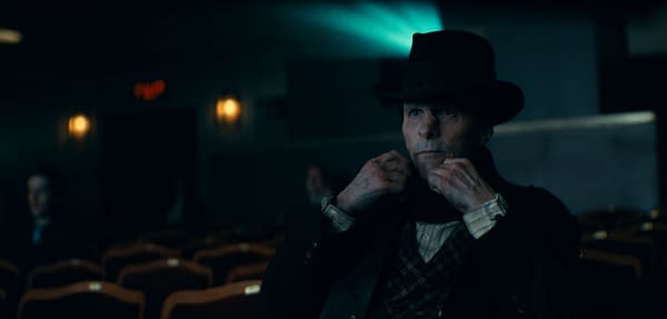 A figure in a darkened movie theater looks intently at the screen, wearing a black hat and adjusting a scarf around their neck. Rows of empty theater seats are visible in front of them, adding to the cinematic atmosphere.