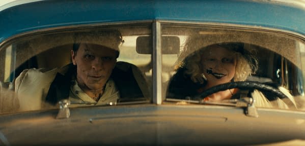 A scene from a film featuring two characters inside an old car; the man has scars on his face and wears a white suit, while the woman, with curly blonde hair and painted face makeup, smiles at the camera. Dust covers the car's interior, contributing to a gritty aesthetic.