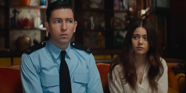 In a scene from 'The Sheep Detectives,' Nicholas Braun portrays Officer Tim Derry, wearing a blue police uniform, sitting next to Molly Gordon as Rebecca Hampstead, who has a concerned expression. The background features a colorful, eclectic decor.