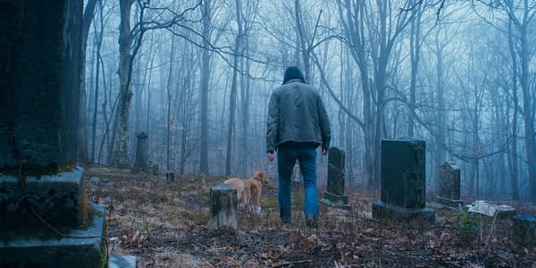 A person wearing a dark jacket walks through a misty cemetery alongside a golden retriever, surrounded by tall, bare trees and gravestones partially obscured by fog.