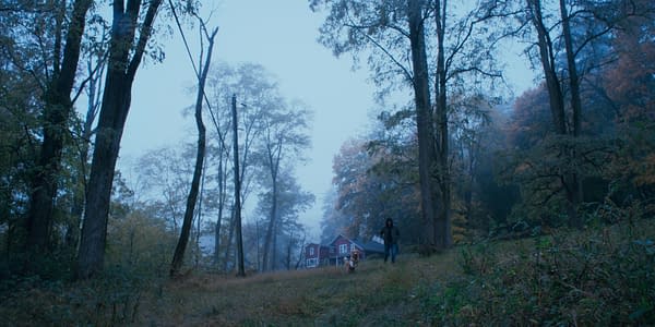 A person walking with a dog on a foggy path in a wooded area, with a red house visible in the background among autumn foliage.