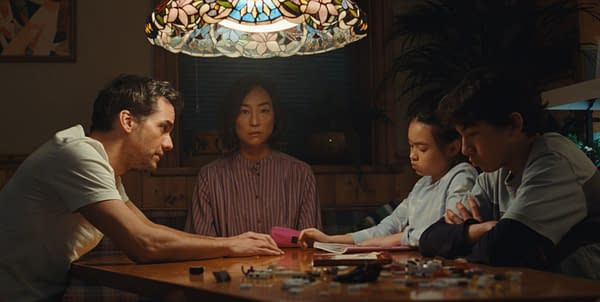 A tense family scene at a dining table, with two adults and two children seated under a stained-glass lamp in a dimly lit house.